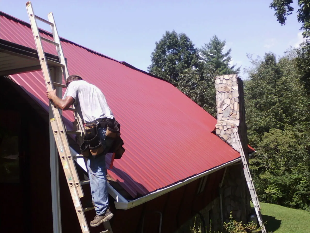 Roofer on a ladder beside a red metal roof and stone chimney — Soffit & Fascia Repair by Roofing Brothers in Wolcott